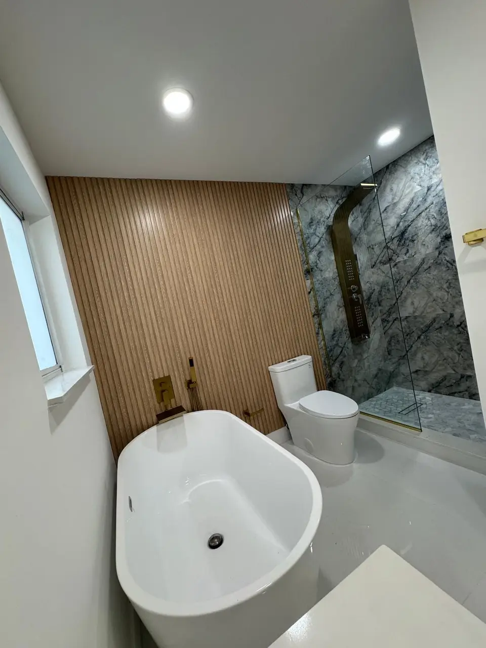 Bathroom with freestanding tub, gold fixtures and dark marble shower behind oak slats